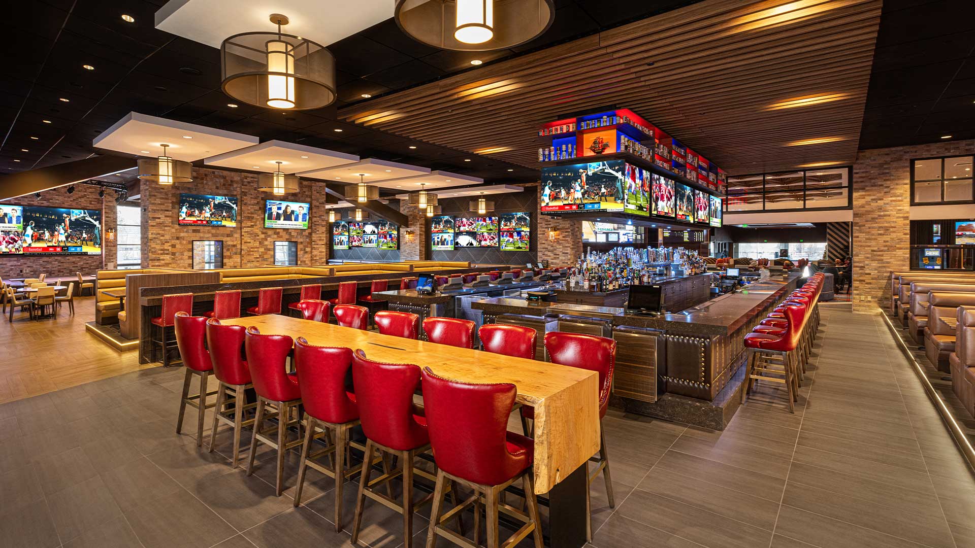 A sports bar with a long wooden table and red leather bar stools, multiple TVs on the walls, and stone wall decor.