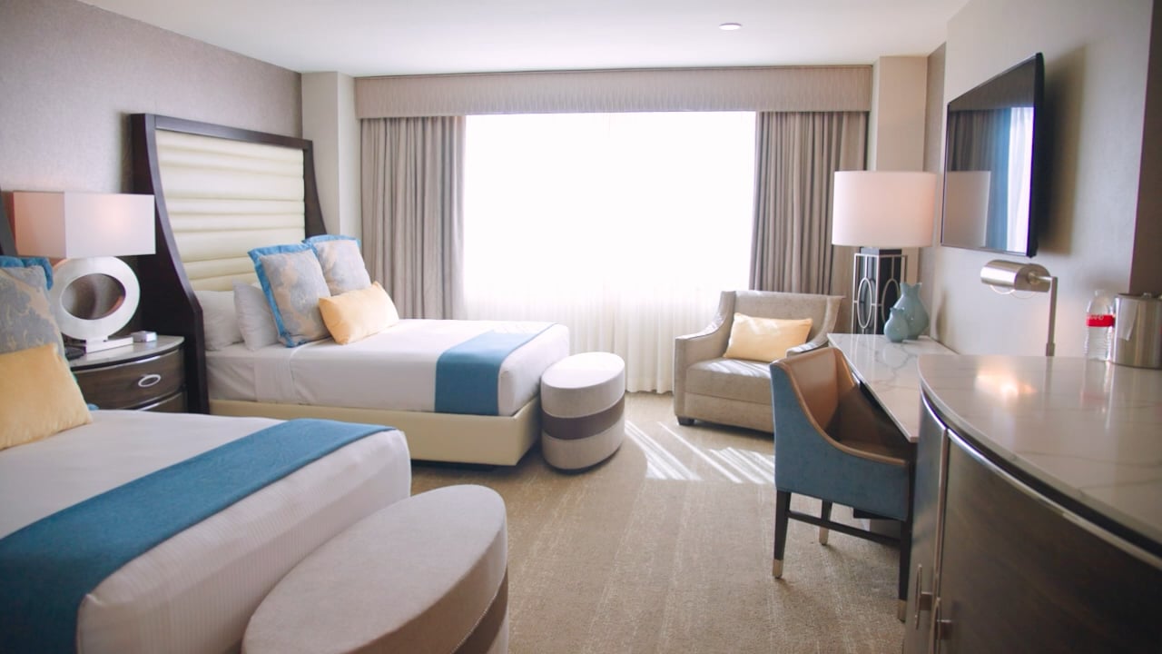 A wide view of a hotel room with two queen beds, a desk, chair, and TV.