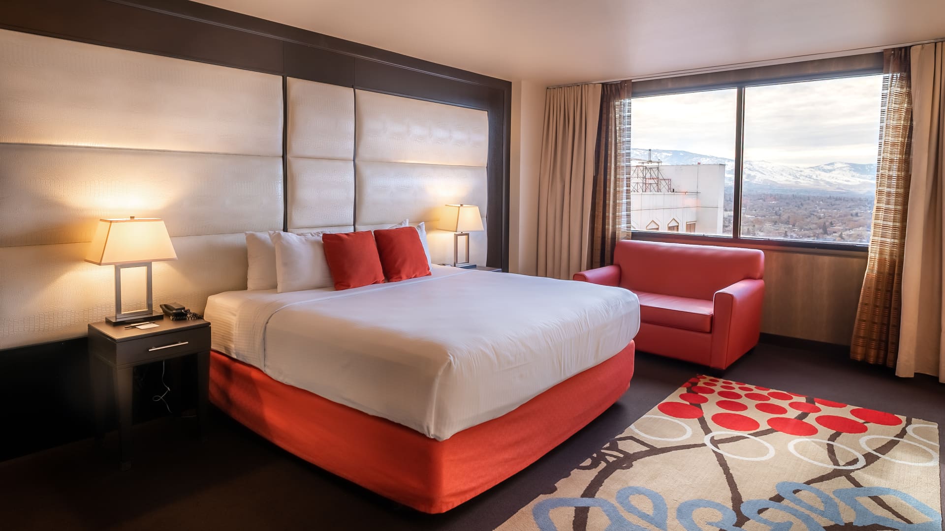 A hotel room with a large bed, red chair, and window with a view.