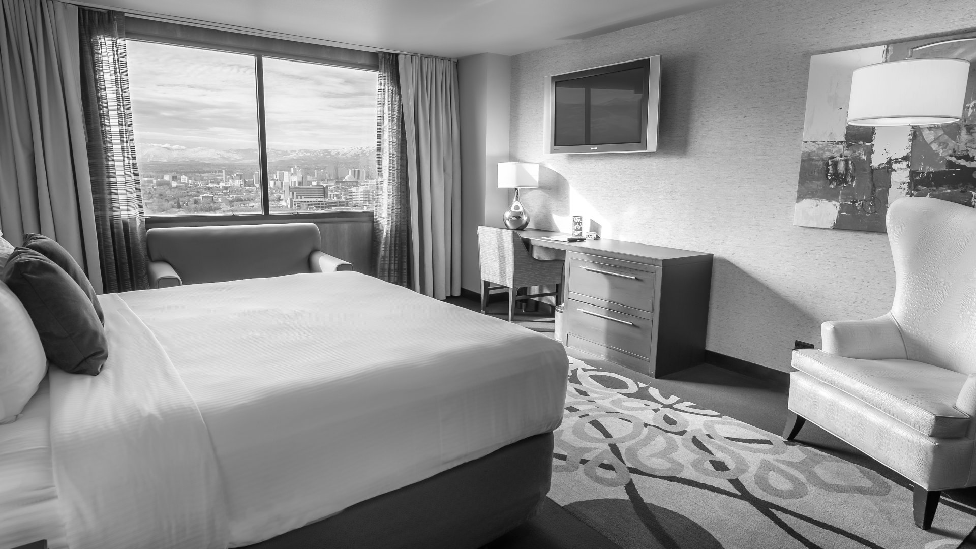 A hotel room with a bed, desk, chair, and TV, overlooking the city.