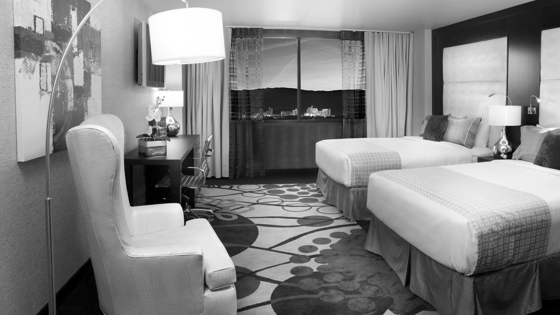 A wide-angle view of a hotel room with two queen beds, a desk, and a chair, looking out onto a city view at dusk.