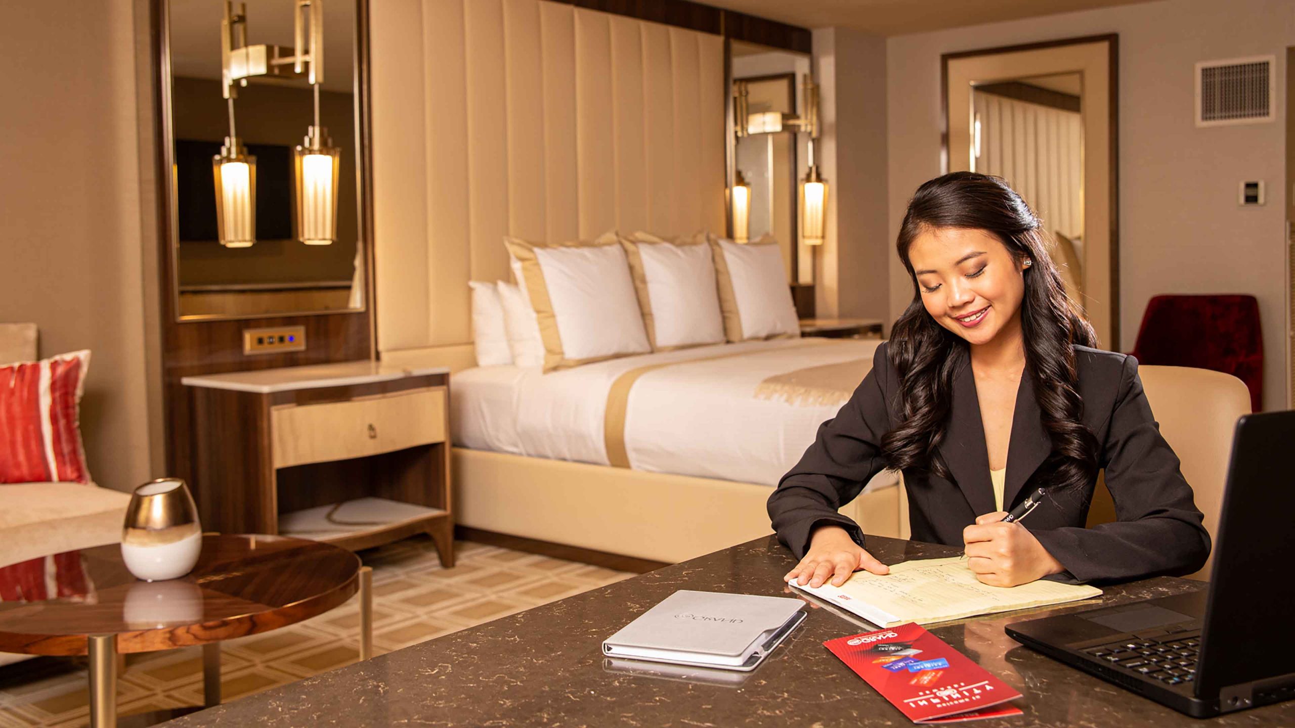 A woman sits at a desk in a hotel room with a bed, sofa, and coffee table.