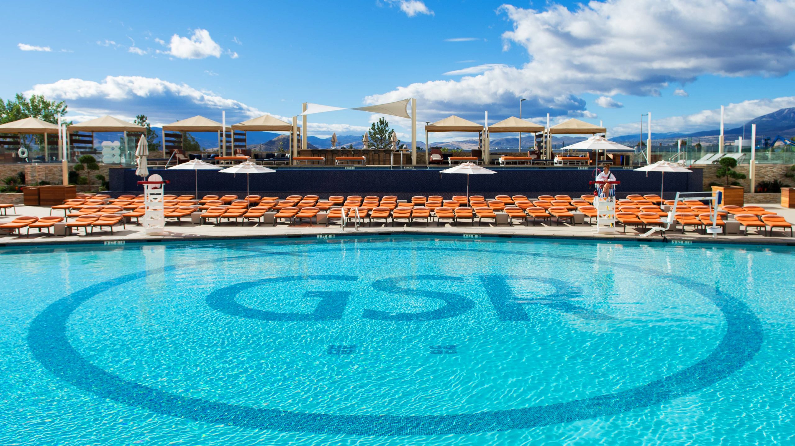 A large outdoor pool with lounge chairs and umbrellas, set against a backdrop of mountains and a blue sky with clouds.