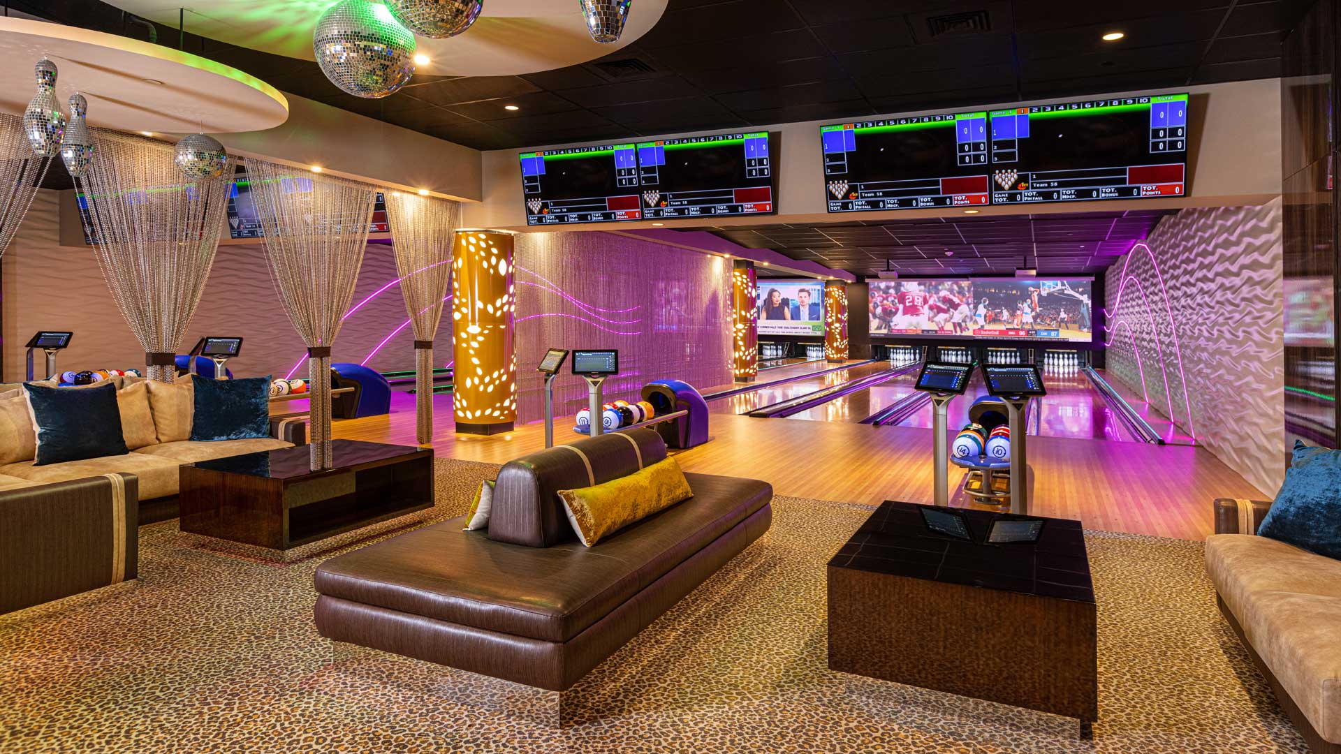 A modern bowling center with a lounge area, featuring a bowling lane, seating areas with couches and coffee tables, disco balls, and large screens displaying scores and a sports game.